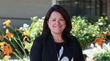Kristi Ferguson, Senior Manager, Communications + Community Relations Woman smiles in front of district building and some greenery/flowers in the garden bed