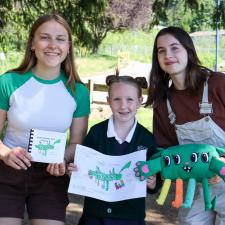 Two female secondary students stand with female elementary student holding their projects