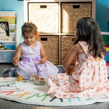 Two young girls sitting on a circular rug playing with a wooden food toy set.