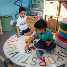 Two young bous playing with wooden cars, sitting on a circular rug on the floor.