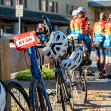 Bikes with names attached to the front handle bars resting along a wooden fence with riders standing in the background out of focus.