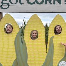 Three elementary students with their heads placed in cut out wooden holes that are panted/designed as corn cobs.
